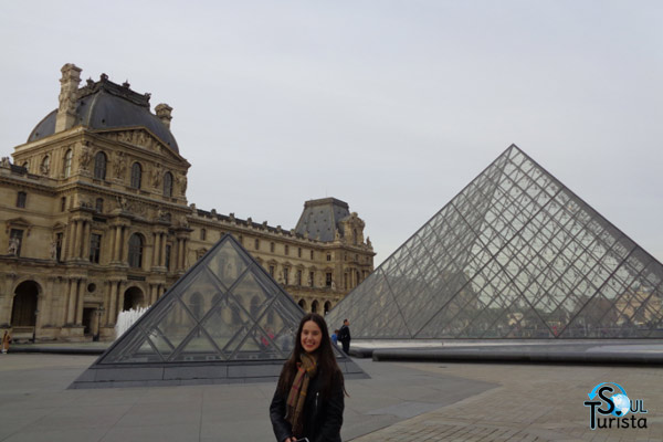 Foto minha na parte externa do Museu do Louvre com as pirâmides