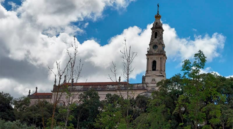 Céu azul, algumas árvores bem verdes que ficam ao redor da igreja e ao fundo está a Basílica Nossa Senhora do Rosário em Fatima Portugal