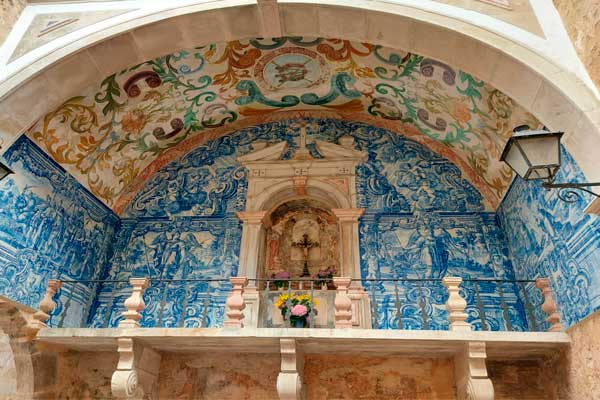 Foto da Ermida da Memória por dentro. Ela é uma capelinha bem pequena, com um crucifixo no alto do seu altar e nas paredes possuem azulejos azuis típicos portugueses