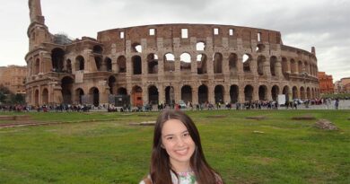 Eu em frente ao Coliseu de Roma. Estou sentada na grade e atrás de mim tem um gramado bem verde e o Coliseu