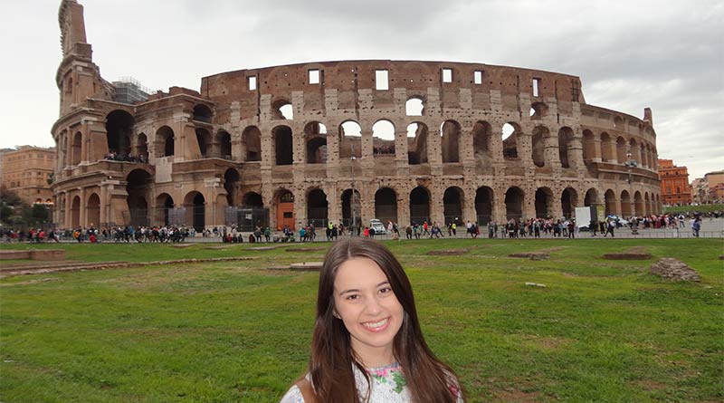 Eu em frente ao Coliseu de Roma. Estou sentada na grade e atrás de mim tem um gramado bem verde e o Coliseu