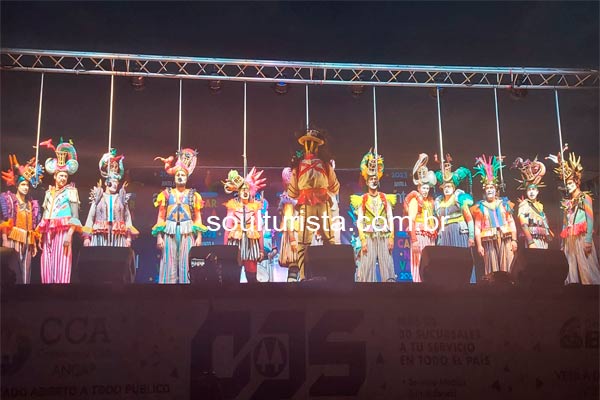 Componentes do Grupo La Venganza de los Utileros apresentando em cima do palco do Tablado do Velodromo com roupas coloridas e chapéus carnavalescos